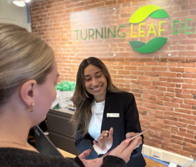 A woman in a black blazer, smiling warmly, assists another woman at a reception desk.